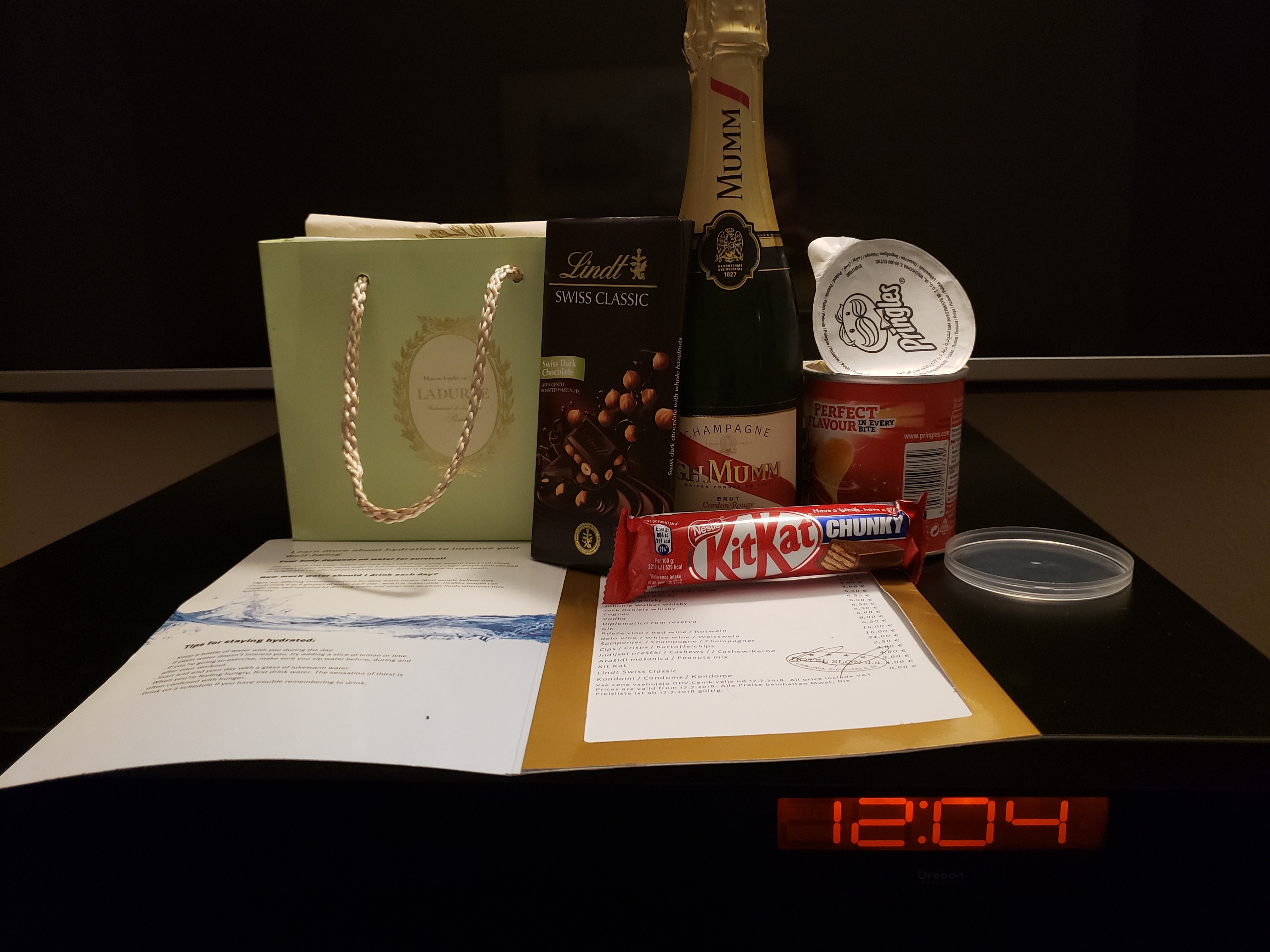 the Laduree sac, a Lindt chocolate bar, a half-bottle of Mumm's champagne, opened bottle of small Pringles, chunky Kit Kat bar all laid out of the mini bar fridge with a digital clock showing the time: 4 minutes past midnight