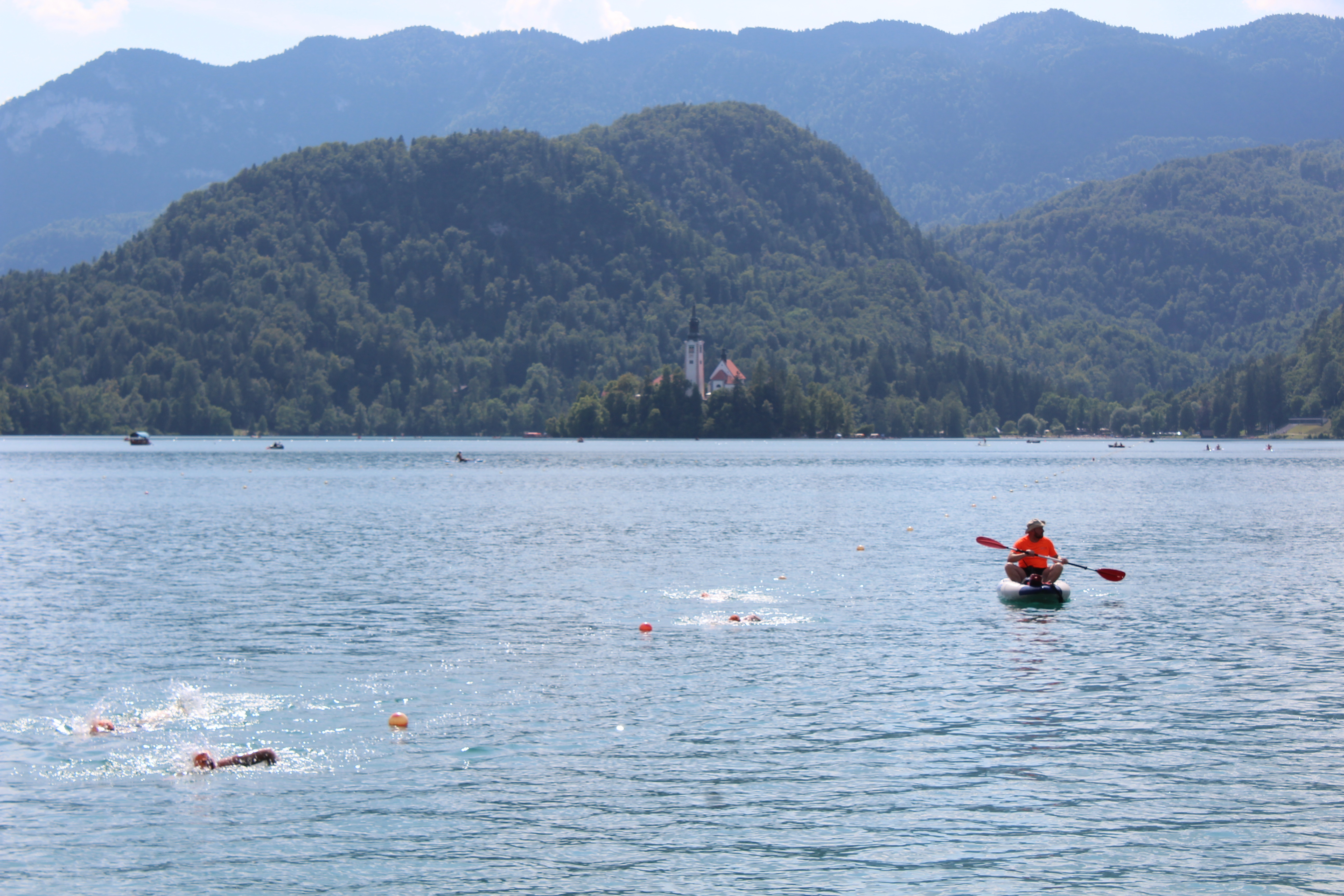 view of the lake with a few swim caps and arms of swimmers, kayaker/guide in a bright orange shirt, mountains in the background and a very small view of the church on the island in the middle of the lake.