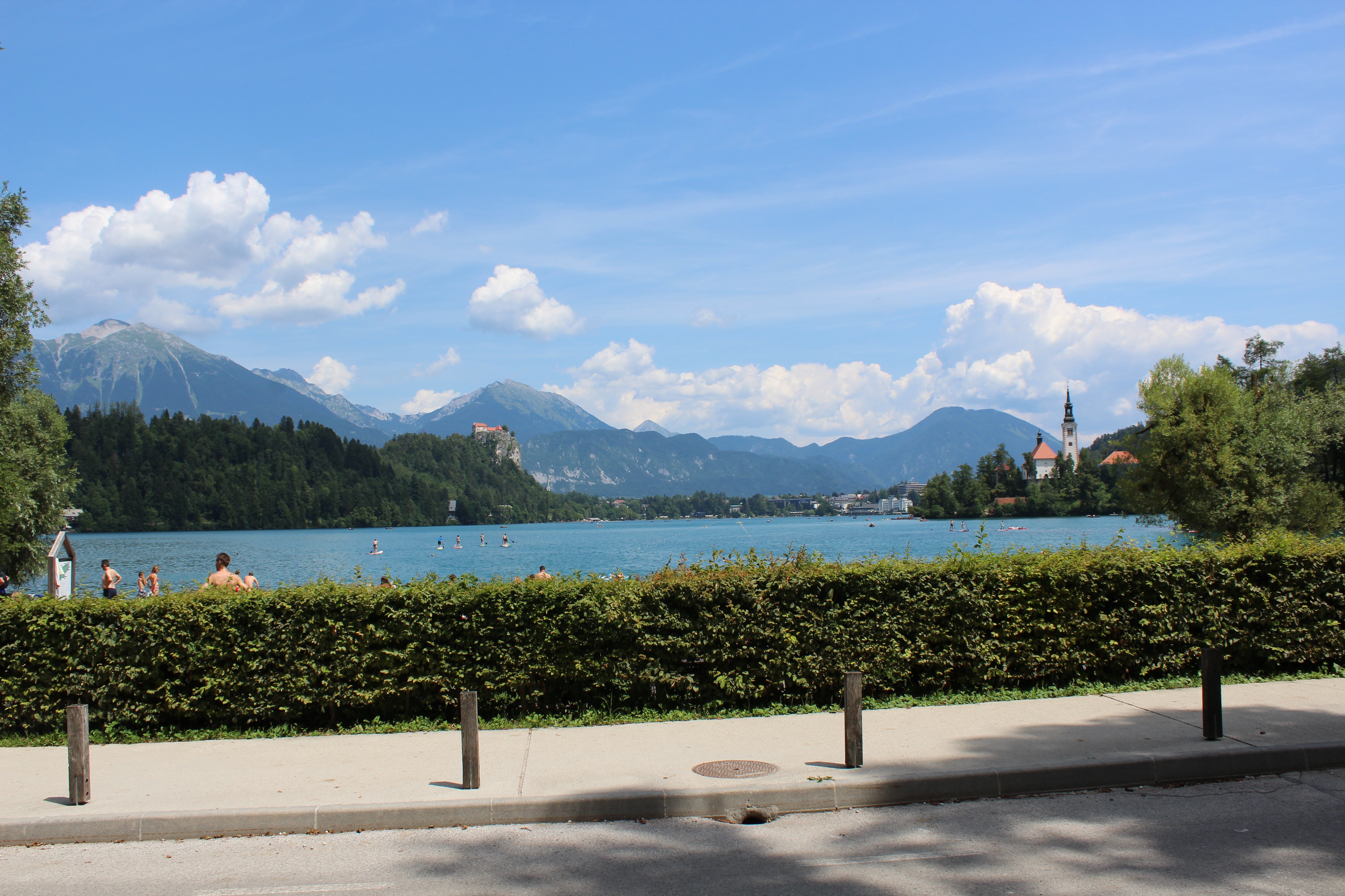 View from the street of the sidewalk, hedges, lake and in the distance the church on the island and mountains in the background