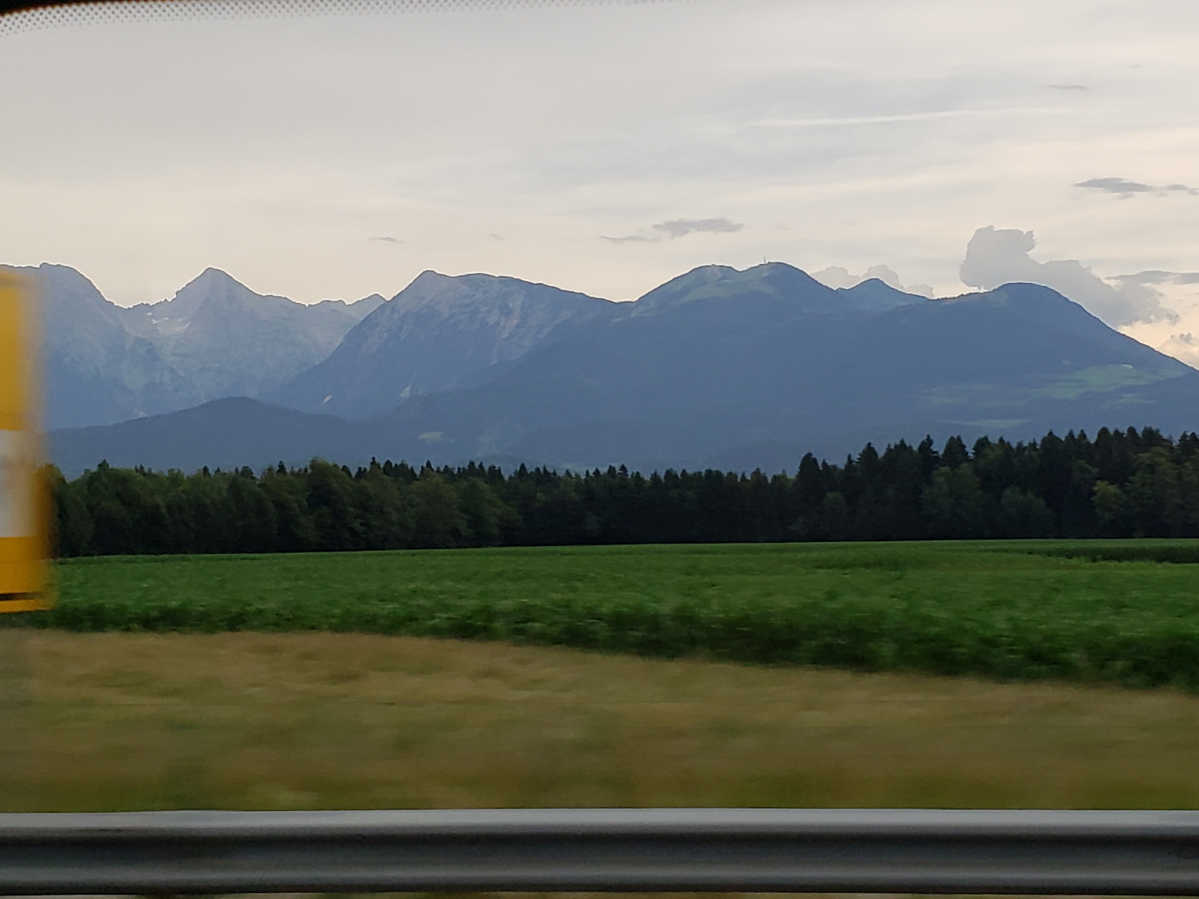view from a van window of yellow grass, then a green crop, beyond that dark green evergreen trees, and in the distance sort of grayed-out you can see mountains that are a mix of tan and dark green, but like there is a film on it making everything a little grey.