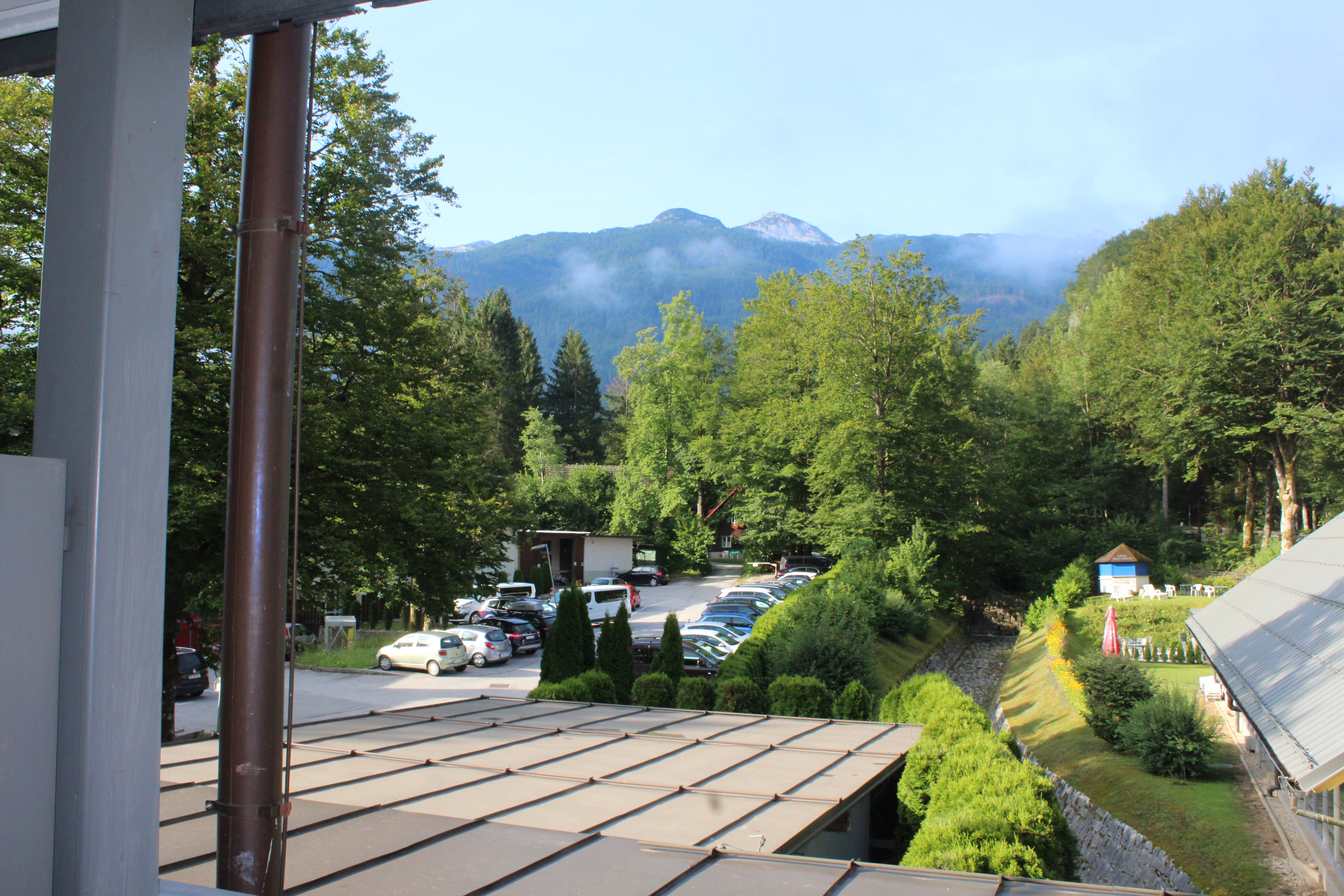 on the bottom of the photo you can see what looks like a metal roof and beyond that structure is a parking lot. There are trees behind the parking lot and beyond that in the distance you can see a mountain with some fog/steam coming off of it
