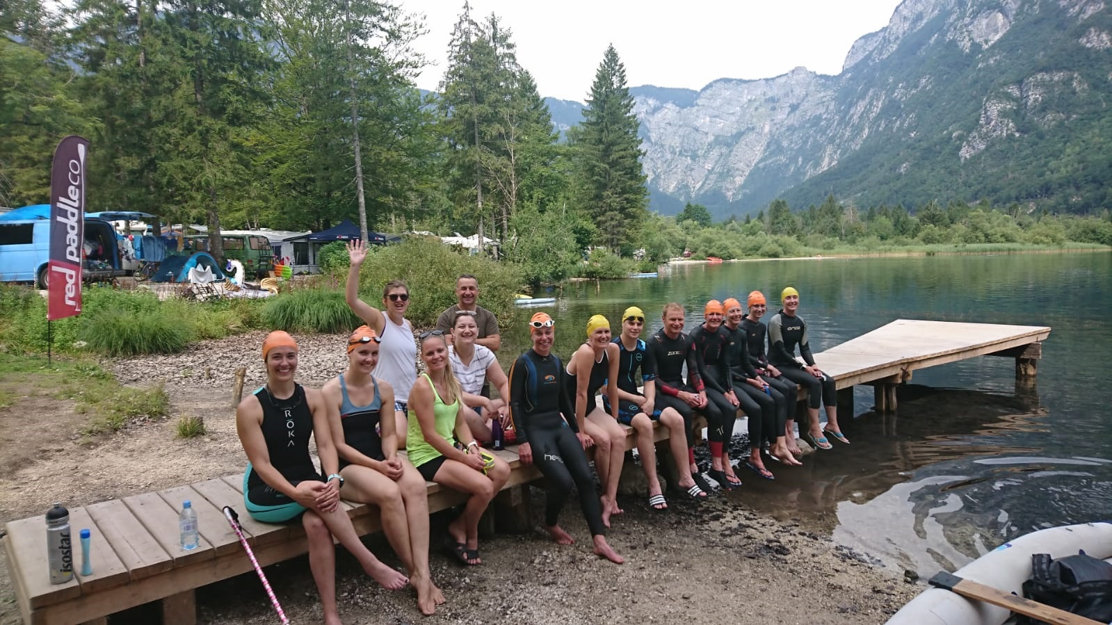 The swim crew decked out in swimsuits and wetsuits sitting on a small wooden dock in lake. Trees and mountain in background