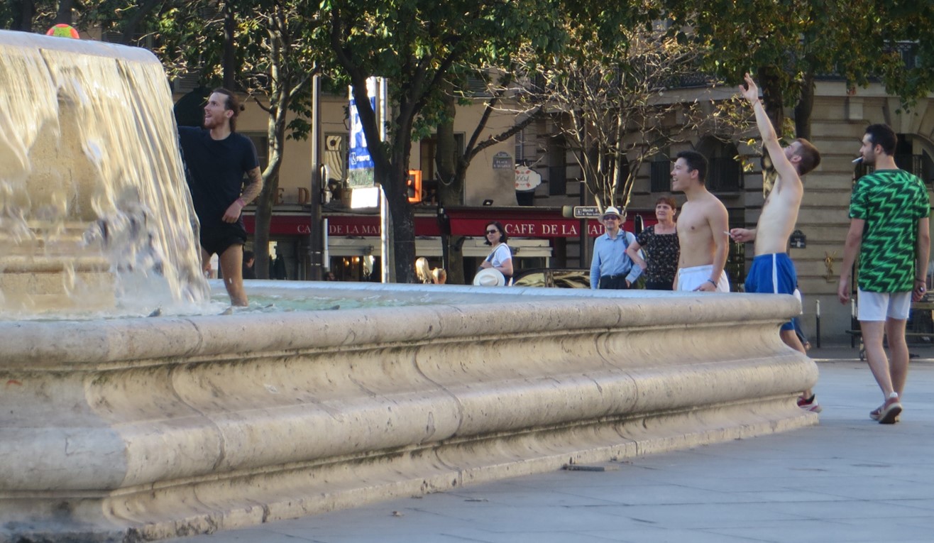 Man dressed in black in the fountain to get his orange and yellow soccer ball. His friends and random people in the square look on.