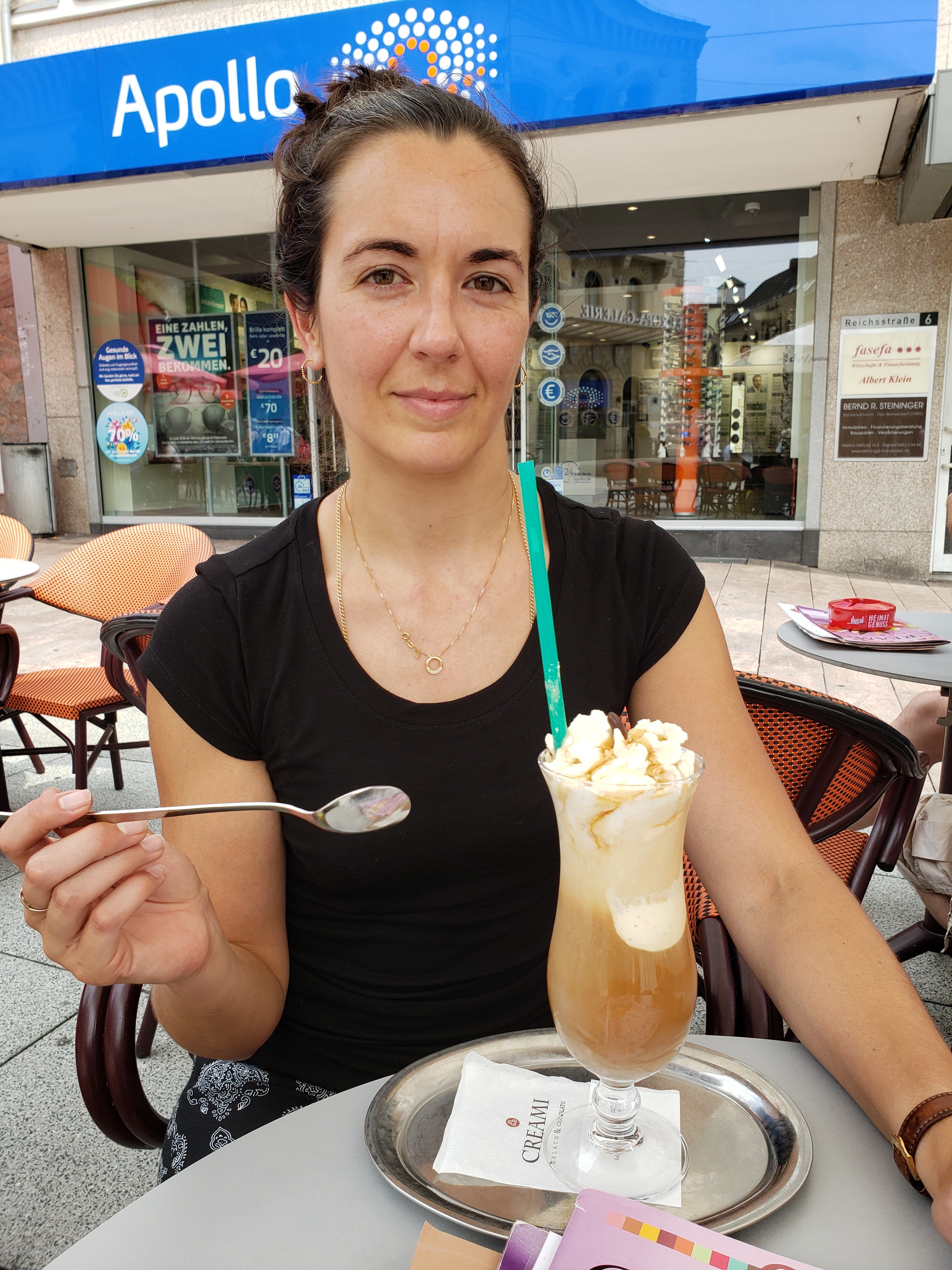 Kerry is wearing a black shirt and is seated at a table with a tall hurricane style glass filled with coffee and ice cream. She is holing a spoon ready to dive into her treat.