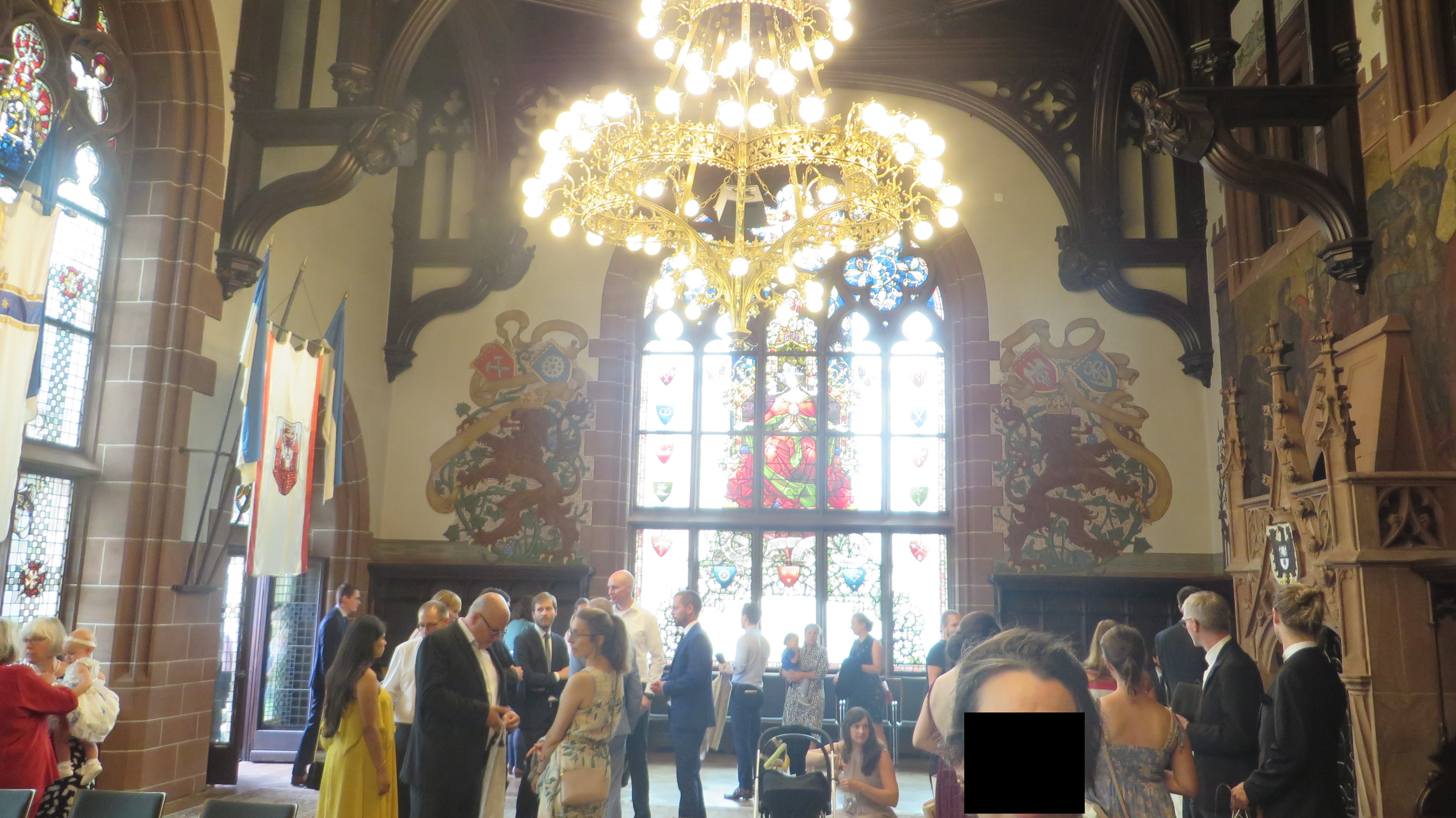 Picture of the inside of the Rathaus where the marriage took place. We see people standing in the room. There is an ornate gold metal 2 tier chandelier. The walls are white with exposed wood and stained glass windows.