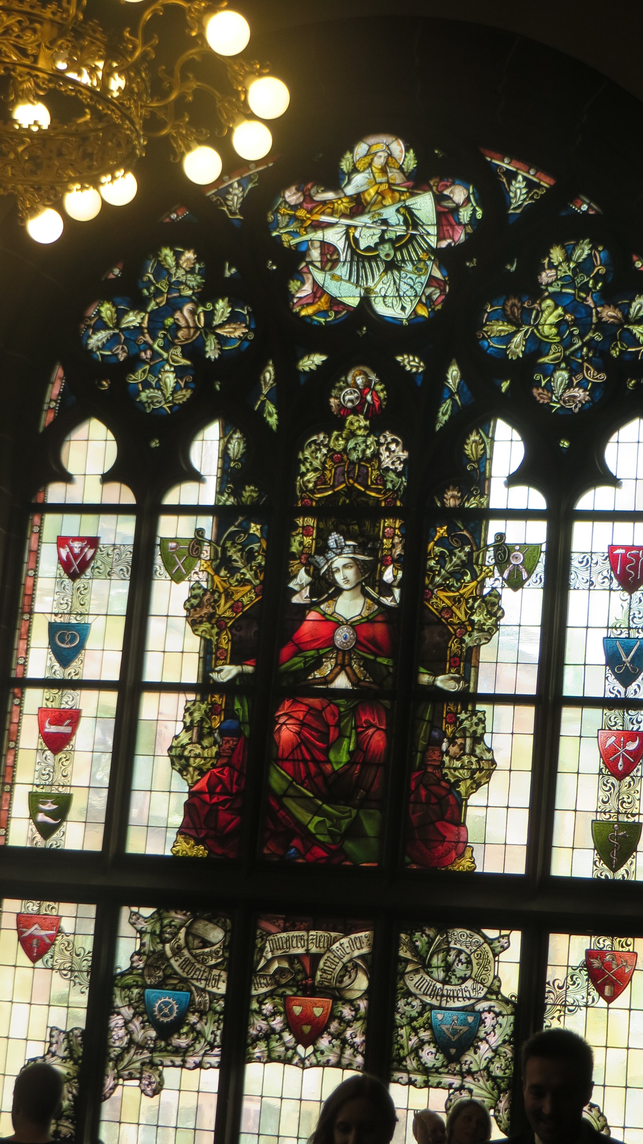 Stained glass window of a woman in red seated on a throne. Different colored/designed shields make a border around her image.