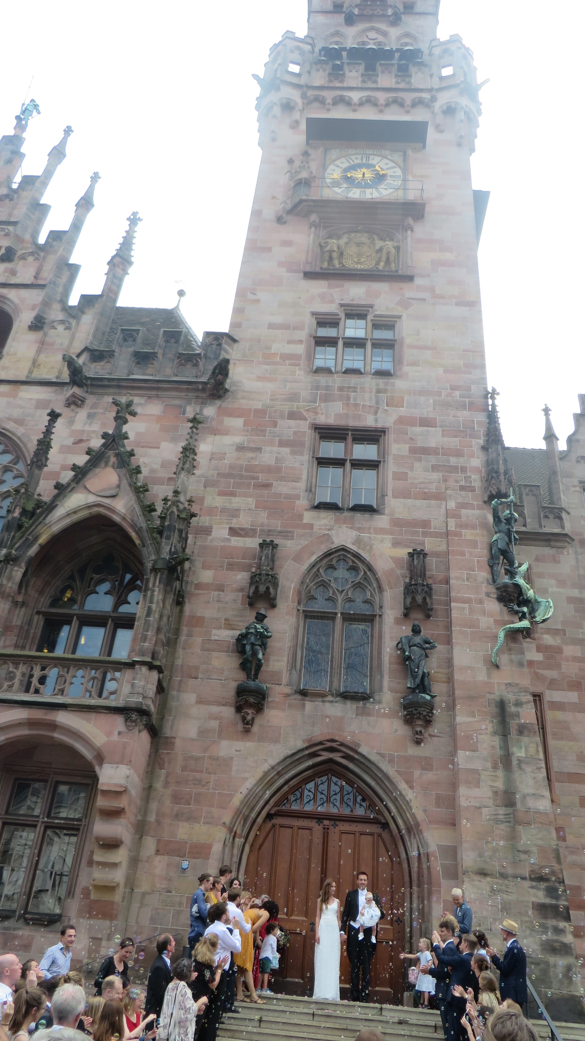 Outside the Rathaus. Standing on the steps are the bride and groom with the groom holding their daughter. The building behind them goes up and forms a clock tower.