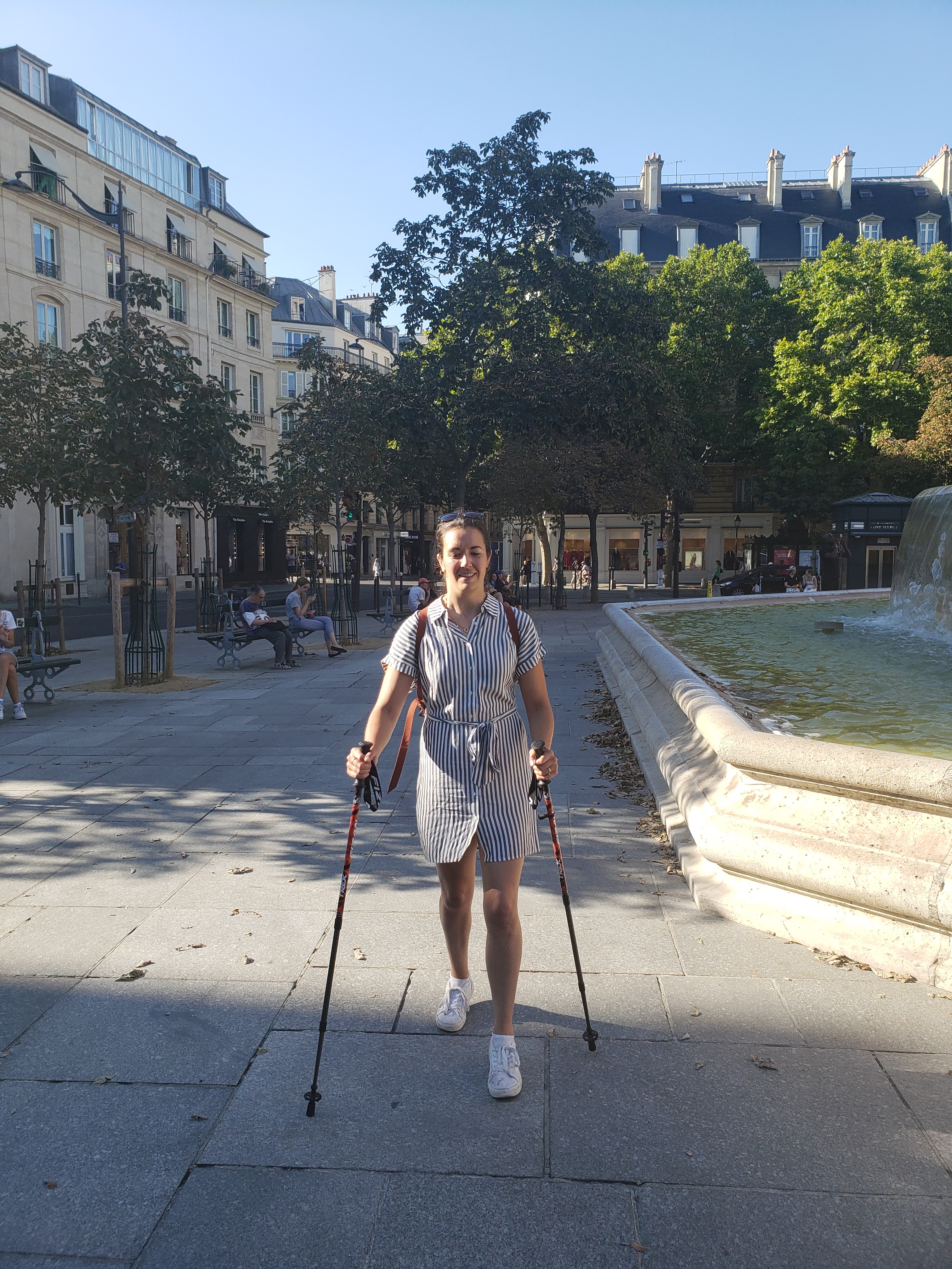 Kerry in a blue and white striped dress, walking with hiking poles in a Paris park square.