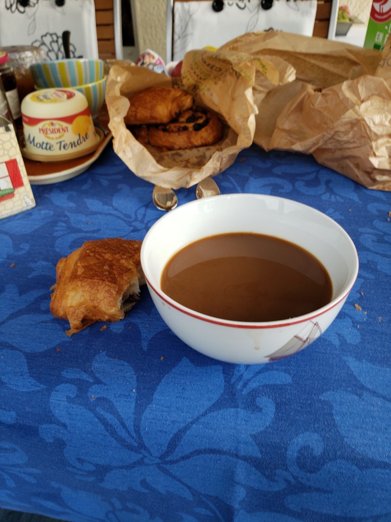 Table with a blue tablecloth. A white bowl filled with coffee, and a half eaten chocolate croissant to the left. In the background is a bag of breakfast pastries and a bucket of butter.