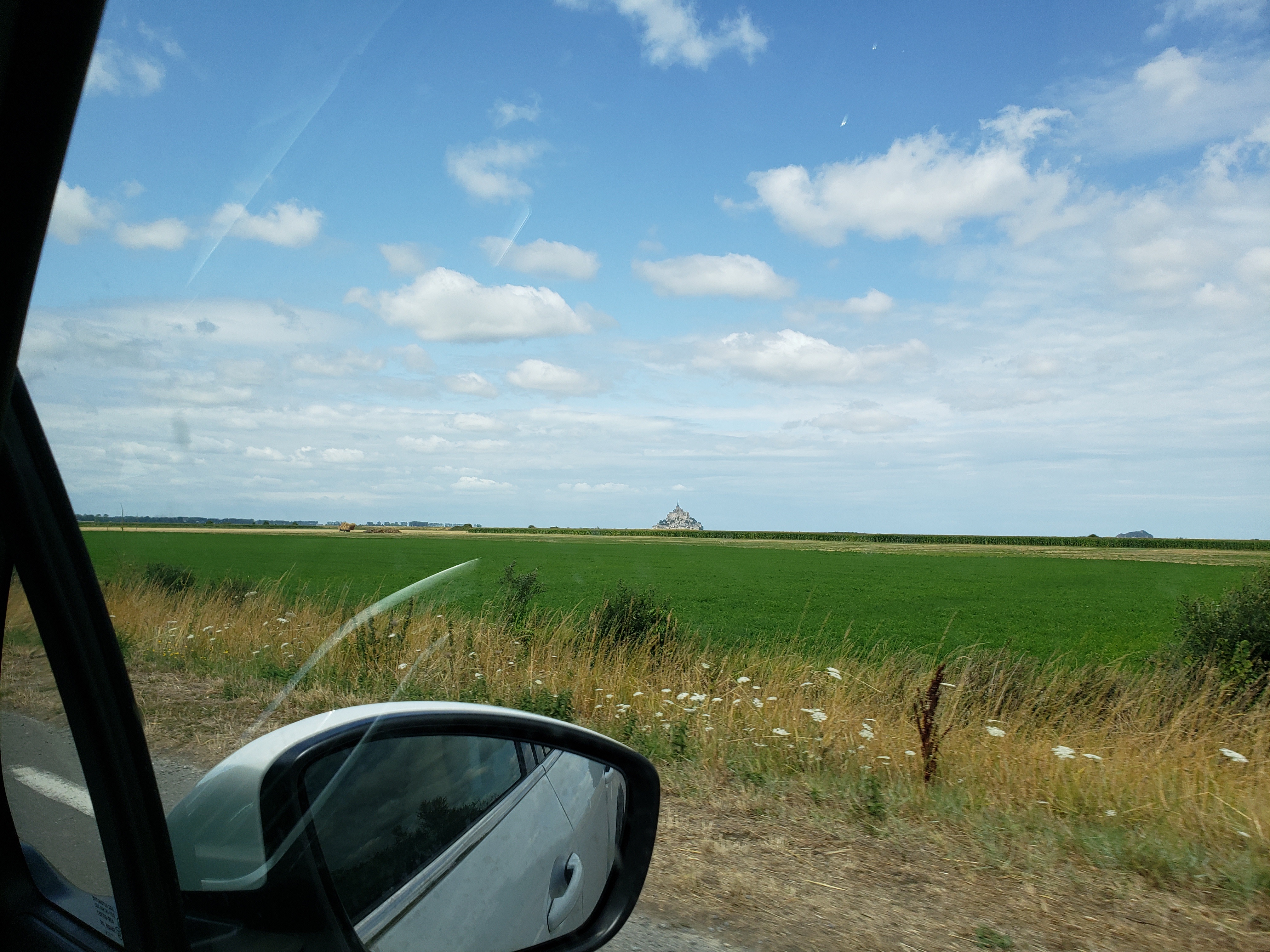 Foreground is the side mirror of the car we were in. Then green fields, clear blue skies with puffs of clouds, and a blob in the middle which is the Mont St. Michel.