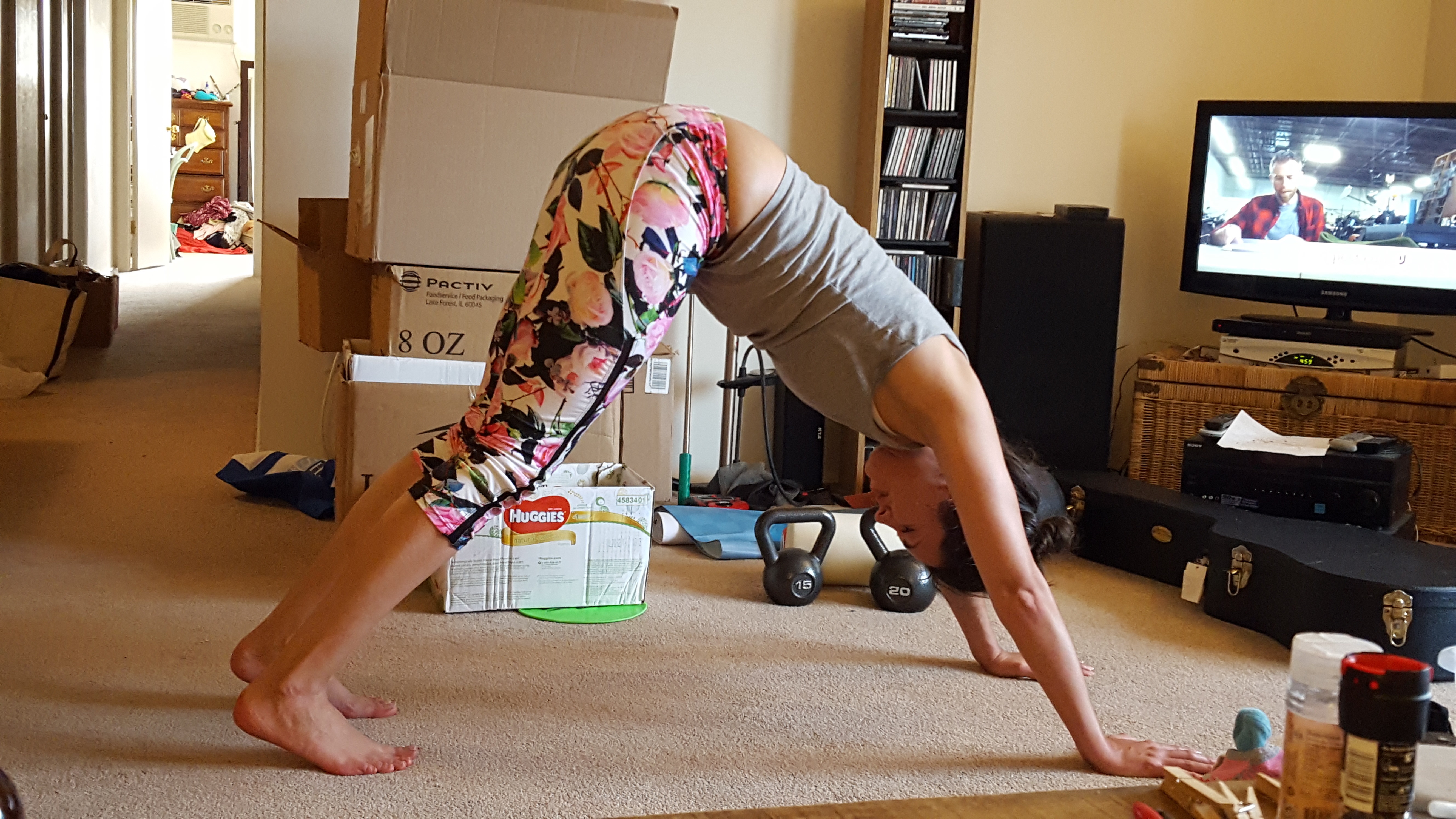 Kerry with a bunch of boxes around her wearing floral leggings and a grey tank top pushing her hips up to the sky forming a triangle with her feet and hands on the floor and hips in the air.