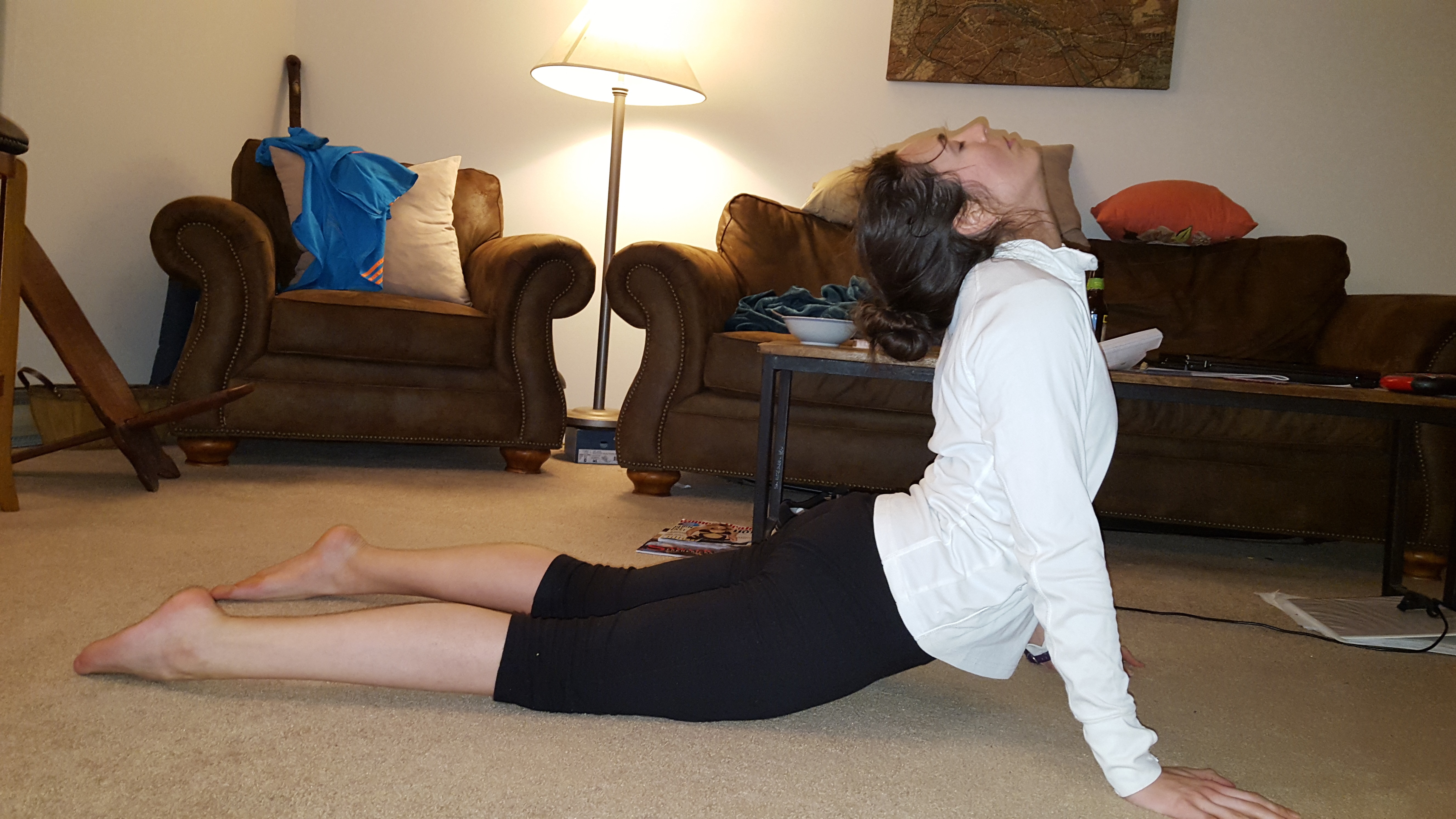 Kerry wearing black leggings and a white longsleeve top. She is doing the updog pose- laying on her stomach she pushed her body upward with her hands.