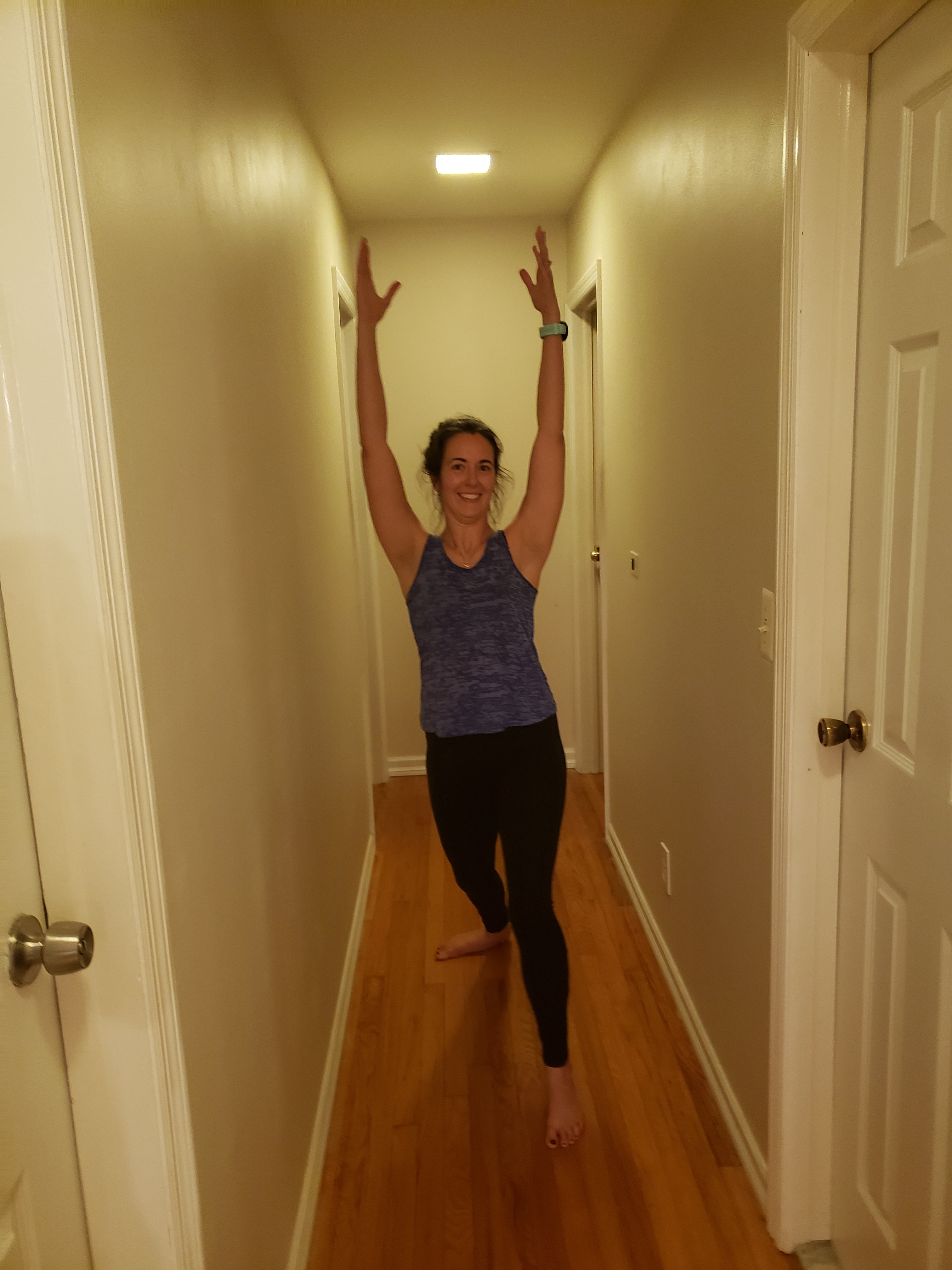Kerry standing in the middle of her hallway with hands up in the air with her left foot forward in a lunge, warrior one position. She is wearing black leggings and a grown tank top.