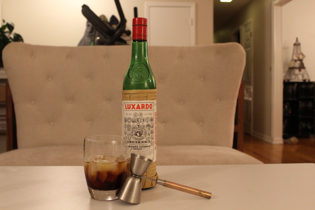 Double old fashioned glass with vermouth, whiskey, and Luxardo in it, bottle of Luxardo, and jigger with a handle.