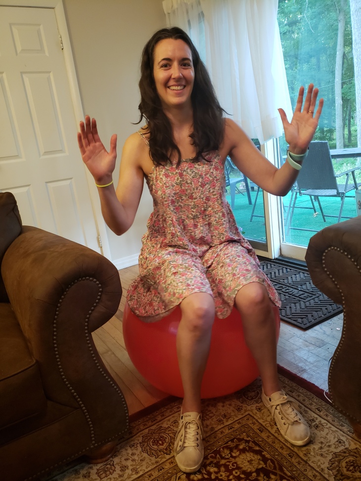 Kerry is wearing a pink floral camisole dress sitting on a red balance ball with sturdy brown couch and chair on either side of her.