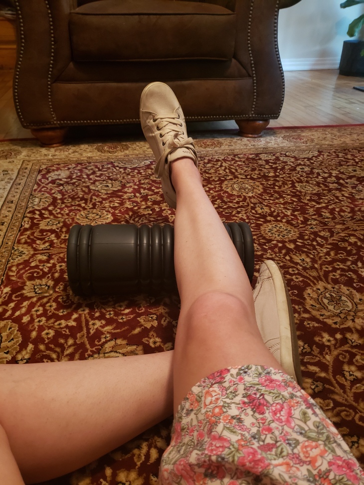 Kerry's right foot is propped up on a black "roller" so she can do ankle exercises.  Couch is in the background and it all is happening on a red and beige carpet.