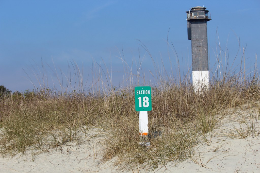 This is a photo of the  green station 18 sign on the left hand side, with sea oats from the dunes in the middle and in the background is the black light house.