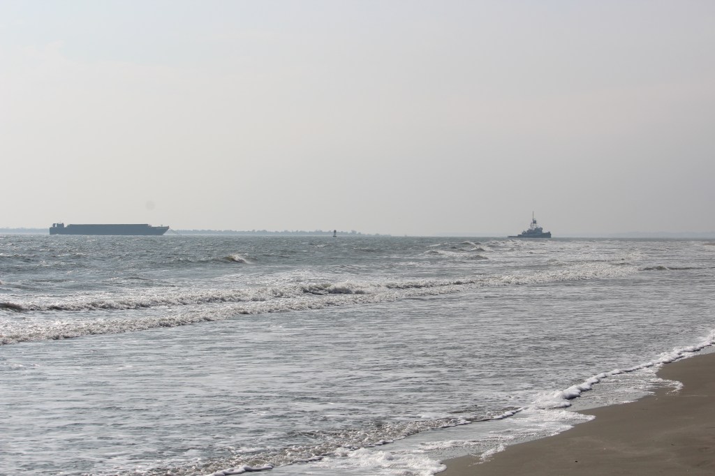 There are a lot of grey tones in this photo. There is a small corner of sand on the right lower corner, with water breaking on the shore.  In the far ground there is a ship on the left with a tug boat pulling it on the right.
