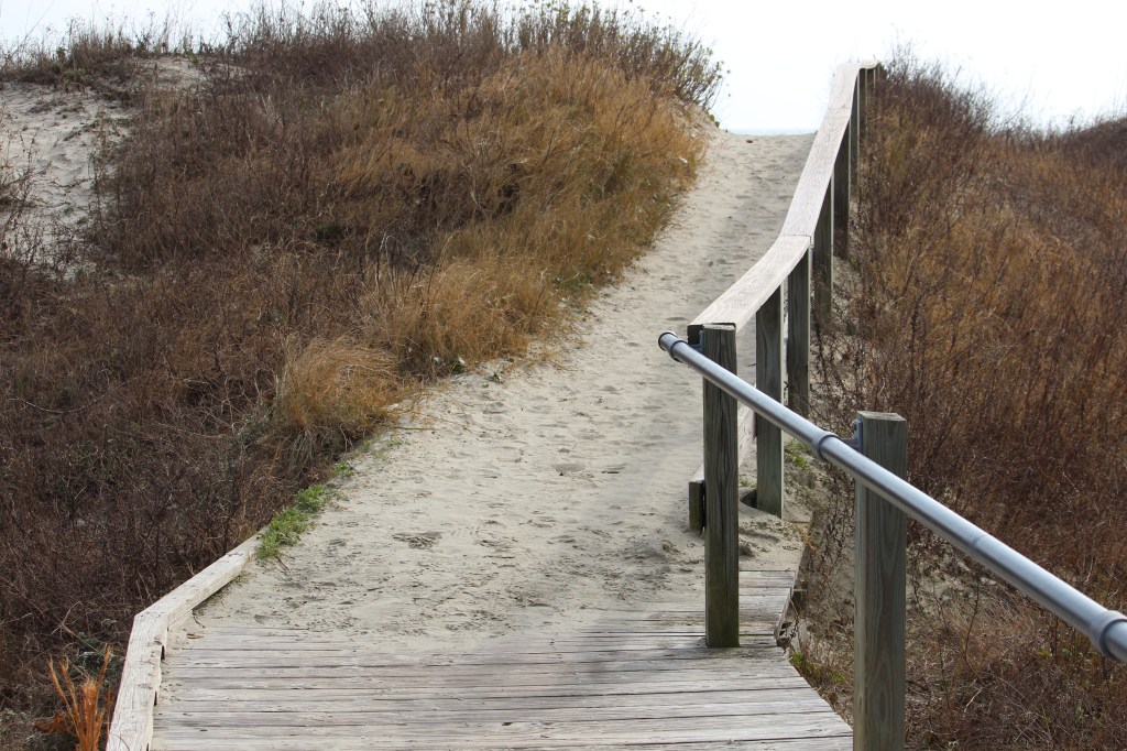 This picture shows the end of the ramp where the railing meets the sand.  It goes uphill  and there is sea grass on either side of the path.